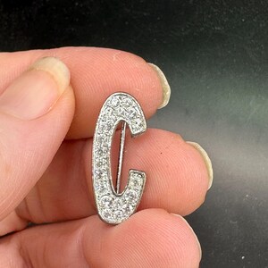 May include: A silver-toned brooch in the shape of the letter "C", encrusted with numerous small, clear crystals. The brooch has a pin on the back for attachment. The brooch is held between two fingers.