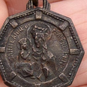 May include: A vintage, octagonal pendant with the image of Sainte Anne de Beaupré. The pendant is a dark, aged metal with embossed lettering and figures. The text "SAINTE ANNE DE BEAUPRE 1927" is visible.