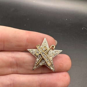 May include: A star-shaped brooch with a silver-tone border and a gold-tone cross and staff design. The star is encrusted with clear rhinestones. The brooch is a vintage accessory.
