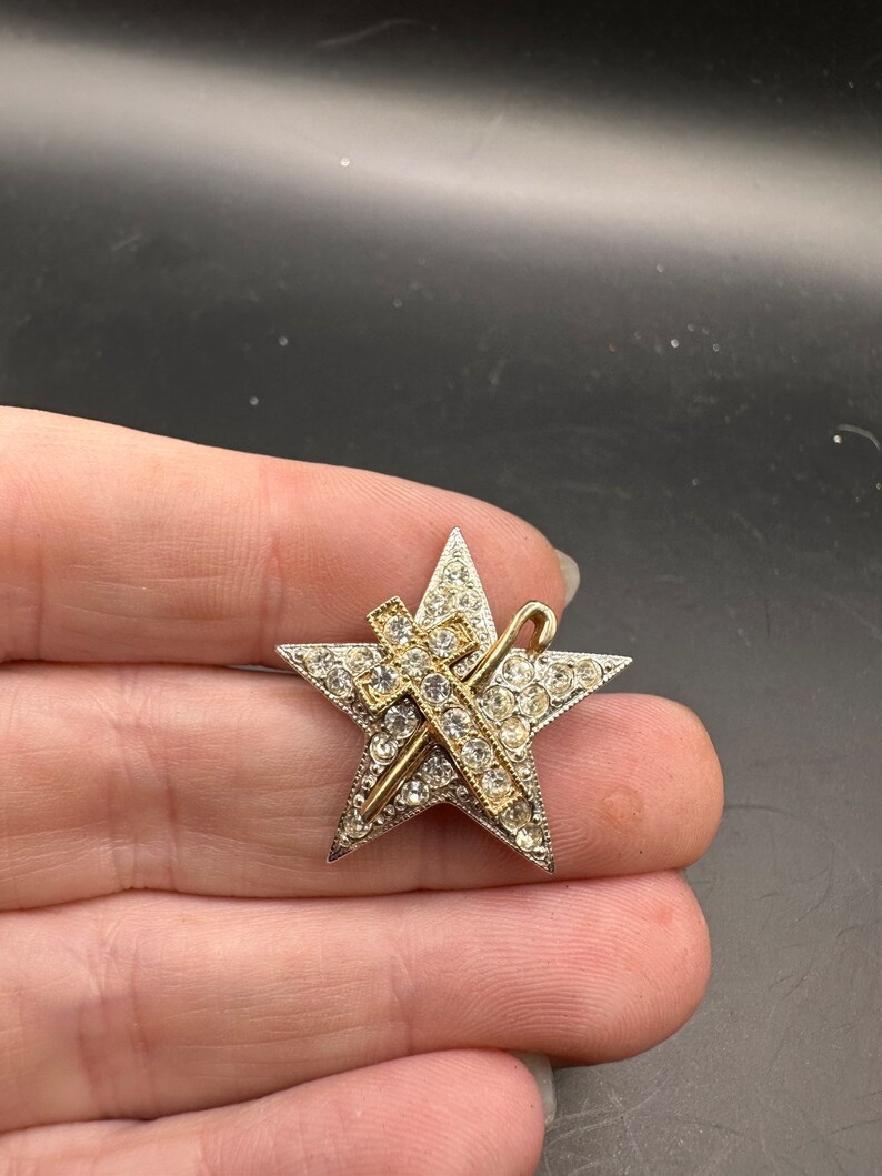 May include: A star-shaped brooch with a silver-tone border and a gold-tone cross and staff design. The star is encrusted with clear rhinestones. The brooch is a vintage accessory.