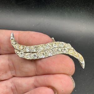 May include: A vintage silver-toned brooch in a curved, leaf-like design. The brooch is encrusted with clear, sparkling rhinestones. The brooch is approximately 3 inches long.