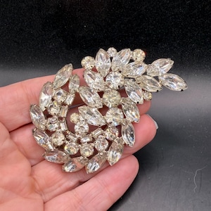 May include: A vintage silver-toned brooch with a floral design. The brooch features numerous clear, sparkling crystals in various shapes and sizes, arranged in a swirling pattern. The crystals catch the light, creating a dazzling effect. A classic piece of jewelry.