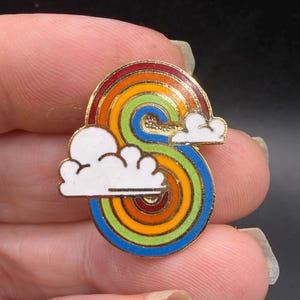 May include: A colorful enamel pin in the shape of the letter "S" with a rainbow design. The rainbow features red, orange, yellow, green, and blue concentric circles. Two white clouds with gold trim are on the left and right sides of the pin.