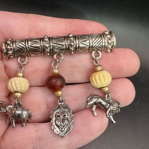 May include: A silver charm bracelet with a silver bar and three dangling charms. The charms are a brown bead, a white bead, and a silver lion head. The bracelet is on a hand.