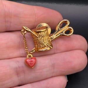 May include: A gold-toned metal brooch featuring a textured spool of thread and a pair of scissors. A small gold chain hangs from the spool, attached to a tiny, smooth, heart-shaped charm with a mottled pink and red enamel finish.