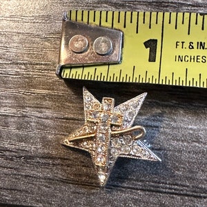 May include: A silver-toned star-shaped brooch with a gold-colored cross overlay, encrusted with clear rhinestones. The brooch is approximately 1 inch in diameter. A metal tape measure is in the background.