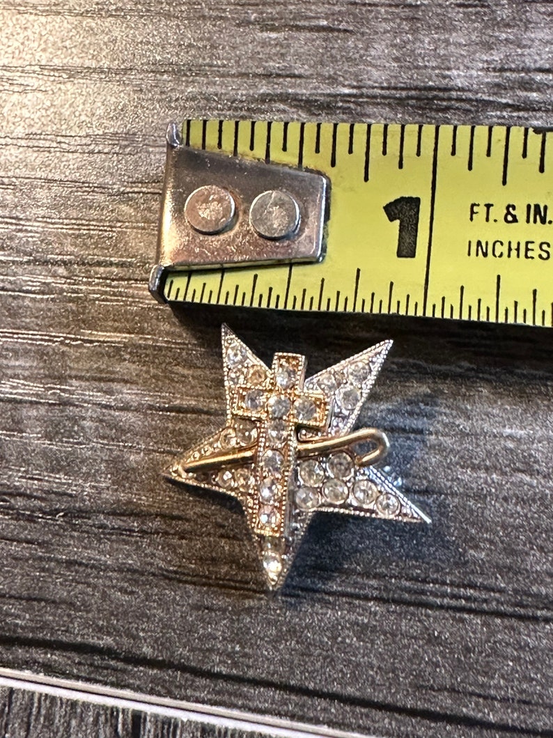 May include: A silver-toned star-shaped brooch with a gold-colored cross overlay, encrusted with clear rhinestones. The brooch is approximately 1 inch in diameter. A metal tape measure is in the background.