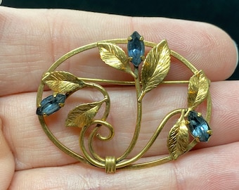 May include: A gold-toned brooch with a floral design featuring three blue gemstones. The brooch is oval-shaped and has a delicate, intricate design.
