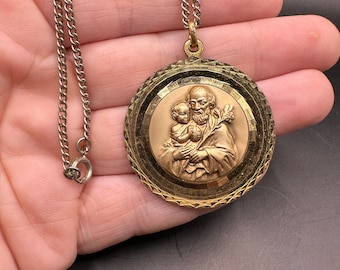 May include: A gold-tone pendant necklace featuring a round medallion with a raised image of Saint Anthony of Padua holding a baby Jesus. The medallion has a textured, patterned border. The necklace is on a silver chain.
