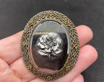 May include: A silver and gold tone brooch with a detailed floral design. The brooch features a raised, silver-toned rose with a black background.