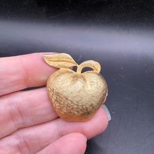 May include: A gold-toned apple-shaped brooch with a textured surface and a leaf. The brooch is likely vintage, with a detailed design. The apple has a stem and a leaf, adding to its realistic appearance. The brooch is a classic accessory.