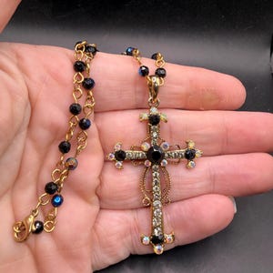 May include: Gold-toned cross pendant necklace with black and iridescent accents. The cross is adorned with black and clear crystals, and the chain features black beads. A decorative piece of jewelry.