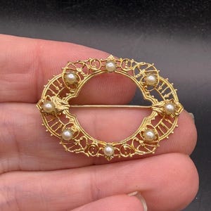 May include: A gold-toned oval brooch with delicate filigree and small pearl accents. The brooch features an openwork design with a pin closure on the back. The jewelry piece is held in a hand.