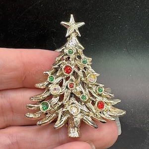May include: A gold-toned Christmas tree brooch with a star on top. The brooch is adorned with red, green, and clear gemstones. The tree has a textured surface and is a festive accessory.