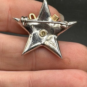 May include: A silver-toned star-shaped brooch with a gold-colored bow and square accent. The brooch has a pin closure on the back and a small, round, yellow-toned stone in the center. The brooch is marked with the letters "DRA".