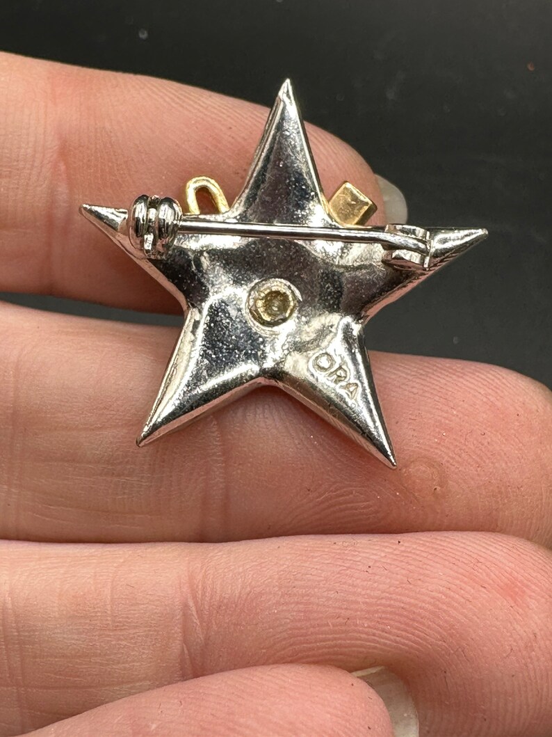 May include: A silver-toned star-shaped brooch with a gold-colored bow and square accent. The brooch has a pin closure on the back and a small, round, yellow-toned stone in the center. The brooch is marked with the letters "DRA".