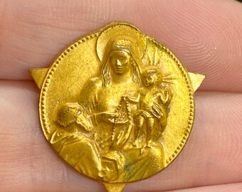 May include: Gold-toned metal religious pendant featuring a relief of the Virgin Mary holding a baby Jesus, with a third figure kneeling in prayer at the bottom. The pendant is circular with a star-shaped frame.