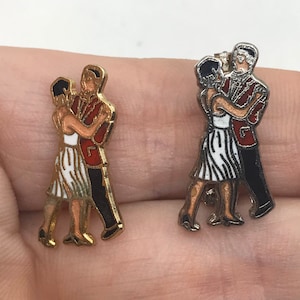 May include: Two gold-tone and silver-tone metal pins depicting a couple dancing. The couple is wearing red, white, and black clothing. The pins are about 1 inch tall.