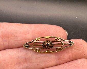 May include: A gold-toned brooch with a delicate design featuring a central black gemstone surrounded by smaller stones. The brooch has a curved shape and intricate details.