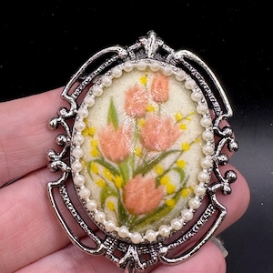 May include: Oval-shaped vintage brooch with a floral design. The brooch features a central image of pink flowers and yellow accents on a cream background, surrounded by faux pearls and a silver-tone metal frame. A classic accessory.