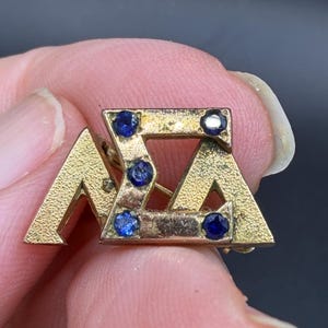May include: A gold-toned pin featuring the Greek letters ΑΣΑ, adorned with small, round, blue gemstones. The pin is held between fingers, showcasing its intricate design and vintage aesthetic. The pin is likely a fraternity or sorority emblem.