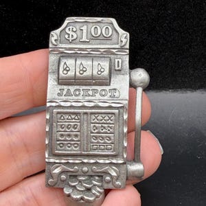 May include: A silver-toned metal belt buckle shaped like a vintage slot machine. The design features the text $100 at the top, three cherry symbols in the display window, the word JACKPOT below, and a pile of coins at the base with a side lever.