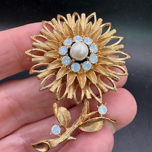May include: A gold tone brooch in the shape of a flower with a faux pearl in the center and clear rhinestones around the pearl. The flower has a stem with leaves and rhinestones.