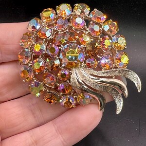 May include: Vintage gold-tone brooch featuring a cluster of iridescent, round rhinestones in shades of orange, yellow, and purple. The brooch has a stylized leaf design on one side. A classic piece of jewelry.