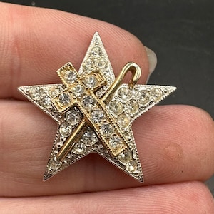 May include: A vintage star-shaped brooch with a silver-tone frame and clear rhinestone accents. A gold-tone cross and staff are overlaid on the star. The brooch is a classic piece of jewelry.