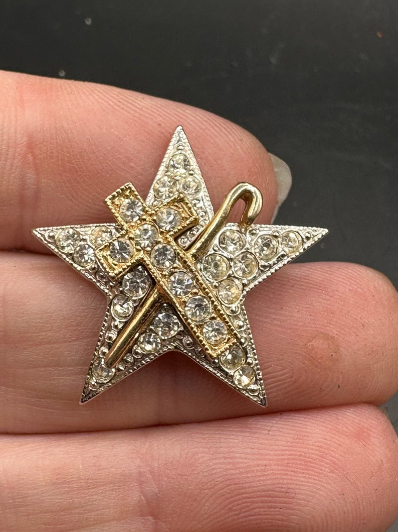 May include: A vintage star-shaped brooch with a silver-tone frame and clear rhinestone accents. A gold-tone cross and staff are overlaid on the star. The brooch is a classic piece of jewelry.