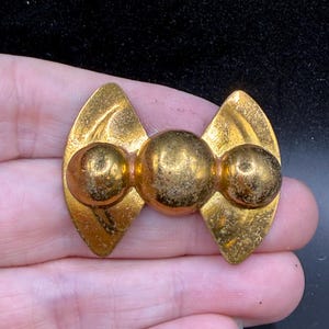 May include: A vintage gold-toned brooch with a bow-tie design. The brooch features three spherical accents in the center and two elongated, leaf-shaped elements on either side. The metal has a slightly aged appearance.