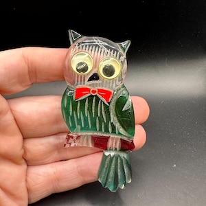 May include: A green and red glass owl brooch with googly eyes and a red bow tie. The owl is perched on a branch and has a silver glitter finish.