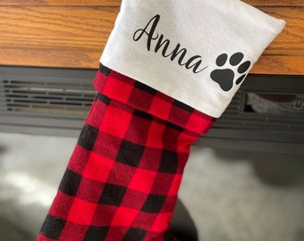 Op de afbeelding: Een kerstsok met een rood-zwart buffelruitpatroon. Het bovenste gedeelte is wit met de naam "Anna" in zwart schrift en een zwarte pootafdruk.
