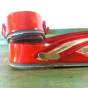 May include: Close-up of a pair of red geta sandals. The wooden platform sandals feature a glossy red finish with gold accents and a red strap. The sandals are on a green surface.