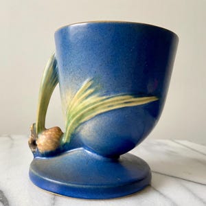 May include: A blue ceramic vase with a unique design. The vase features a bulbous base and a curved handle. The handle is adorned with a tan and green floral motif. The vase is approximately 6 inches tall.