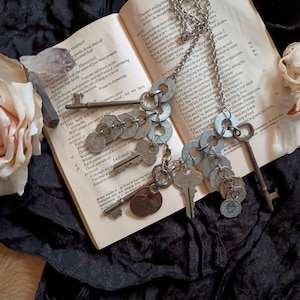 May include: A silver-toned necklace featuring keys, washers, and coins, resting on an open book with text. The necklace has a variety of key shapes and sizes, creating a unique, vintage aesthetic.