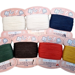 May include: A collection of spools of embroidery thread in various colors, including white, navy blue, green, brown, red, and yellow. Each spool is labeled with Japanese text and a color number.