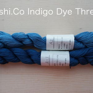 May include: Two skeins of indigo dye thread, one a lighter blue and one a darker blue. The thread is wound around itself and the labels are visible. The labels are in Japanese and say "A-1" and "A-2".