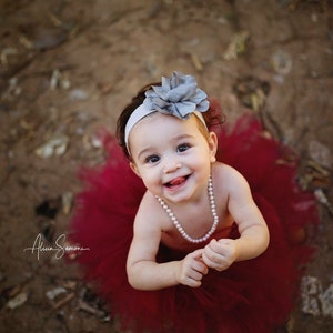 May include: A smiling baby girl wearing a red tulle skirt, a white headband with a gray flower, and a pearl necklace.