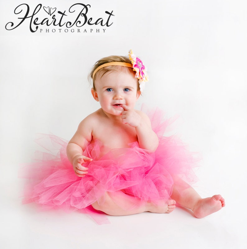 K&ouml;nnte beinhalten: Ein kleines M&auml;dchen tr&auml;gt einen rosa T&uuml;ll-Tutu und ein gelbes Stirnband mit einer Blume darauf. Das Baby sitzt auf wei&szlig;em Hintergrund und hat den Finger im Mund.