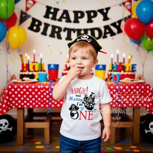 A young child wearing a pirate hat and a white t-shirt with a pirate design is standing in front of a birthday party setup with balloons, a cake, and other decorations.