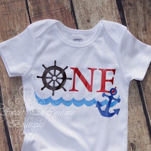 A white baby onesie with a nautical design, including a ship's wheel, anchor, and waves, printed on it.