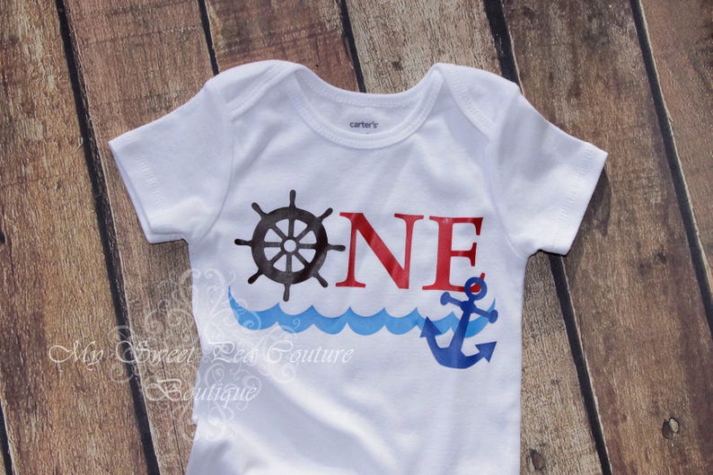 A white baby onesie with a nautical design, including a ship's wheel, anchor, and waves, printed on it.