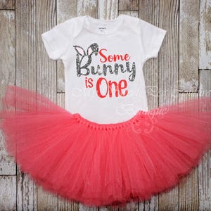 A white t-shirt with the text "Some Bunny is One" printed on it, along with a pink tutu skirt, both placed on a wooden background.