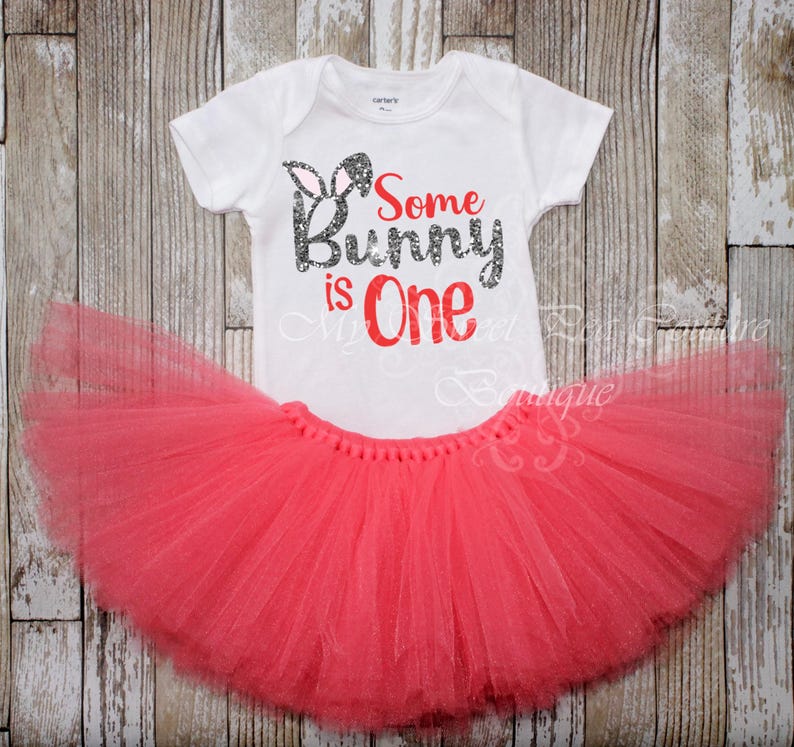 A white t-shirt with the text "Some Bunny is One" printed on it, along with a pink tutu skirt, both placed on a wooden background.