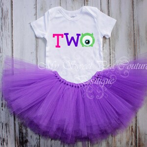 A white t-shirt with a colorful "TWO" design and a purple tutu skirt are displayed on a wooden surface.