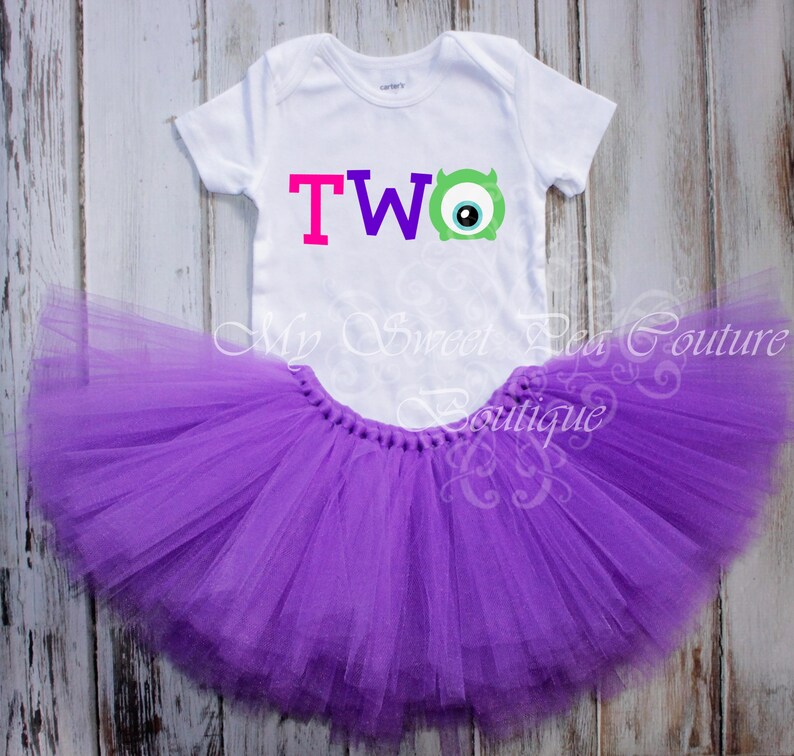A white t-shirt with a colorful "TWO" design and a purple tutu skirt are displayed on a wooden surface.