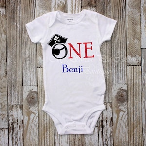A white onesie with a pirate hat and the text "ONE Benji" printed on it, laid out on a wooden surface.