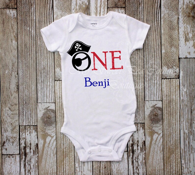 A white onesie with a pirate hat and the text "ONE Benji" printed on it, laid out on a wooden surface.