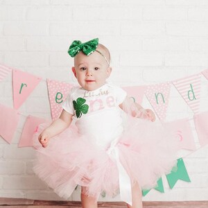 A baby girl wearing a white outfit with a green shamrock and a sparkly green bow stands in front of a white brick wall with pink and green banners.